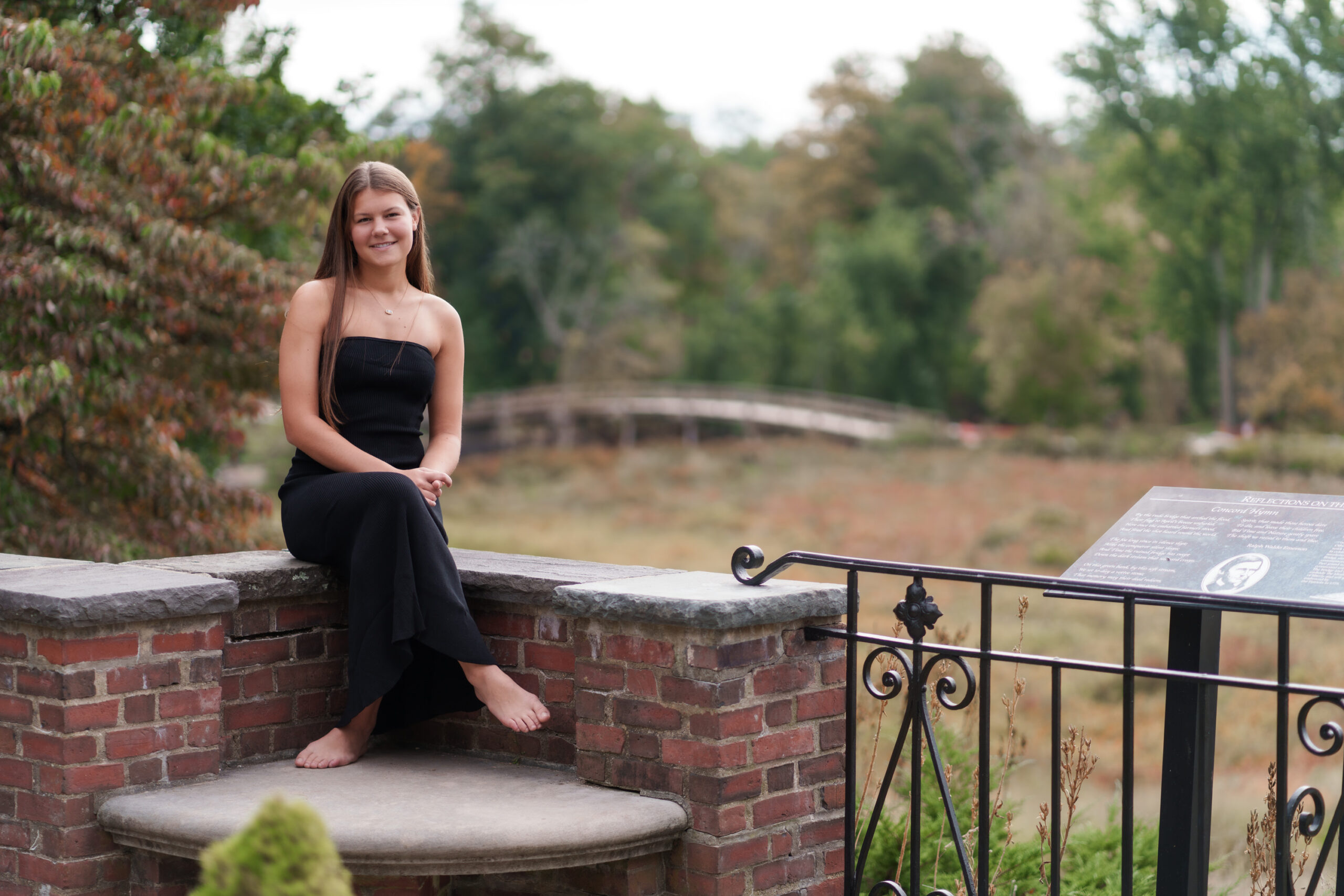 senior portrait of a girl in a long black dress at the north bridge in concord, ma. she's sitting on a stone wall and there is a beautiful meadow and the north bridge in the background. photographed by chiha portraits.
